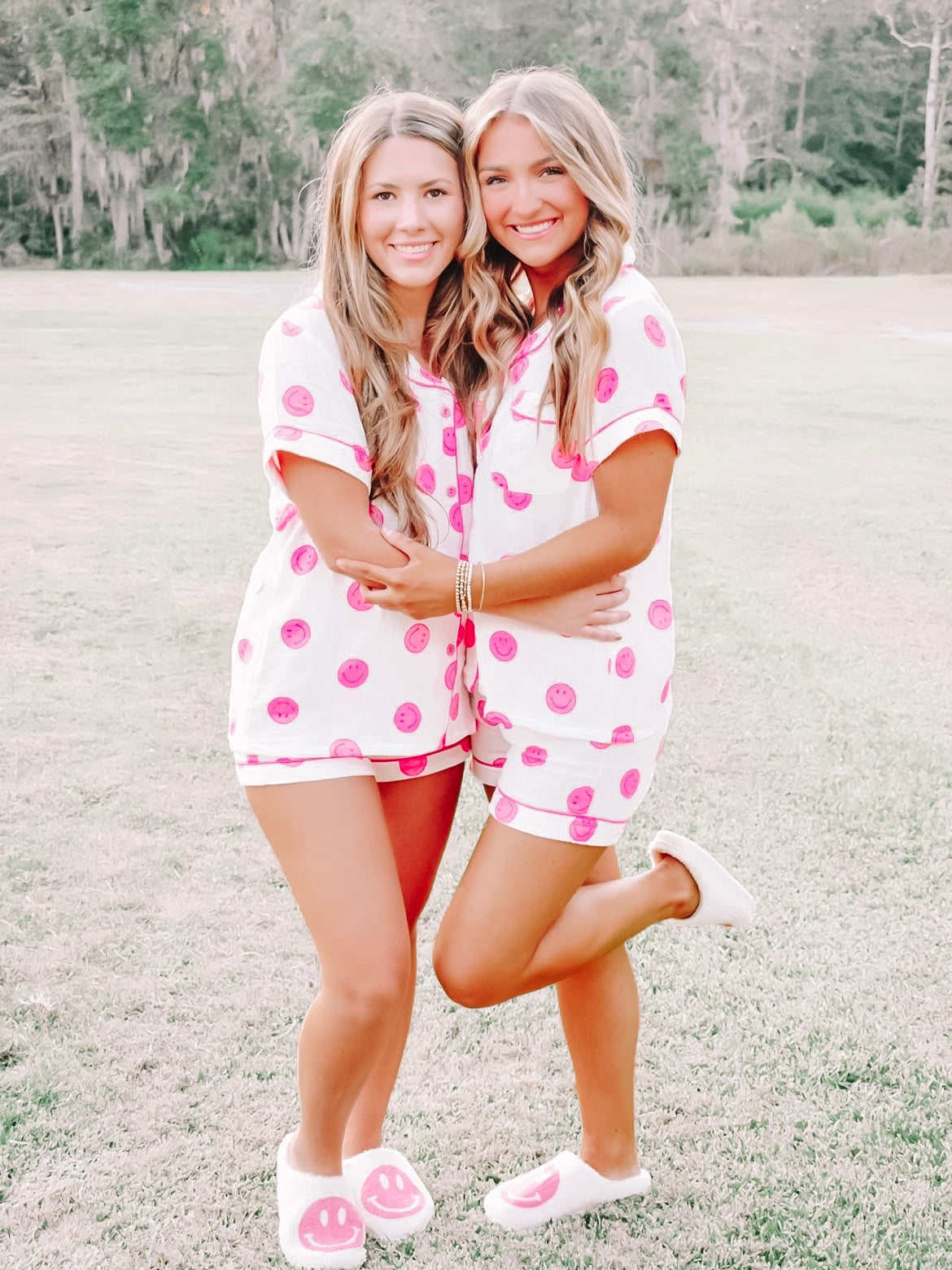 Two women in matching pink smiley face pajama shorts and slippers, outdoors on a grassy field.