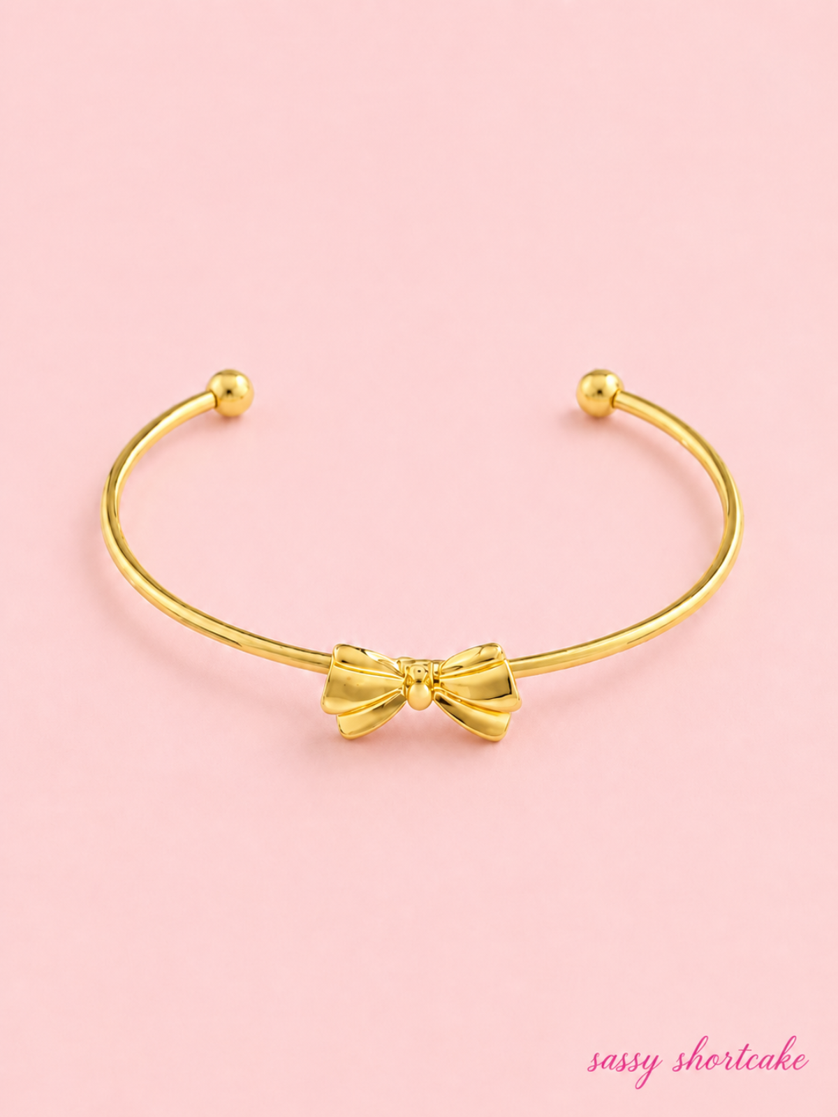 Golden Girl Bow Bracelet from Sassy Shortcake: Gold open cuff bracelet with a dainty bow detail on a light pink background