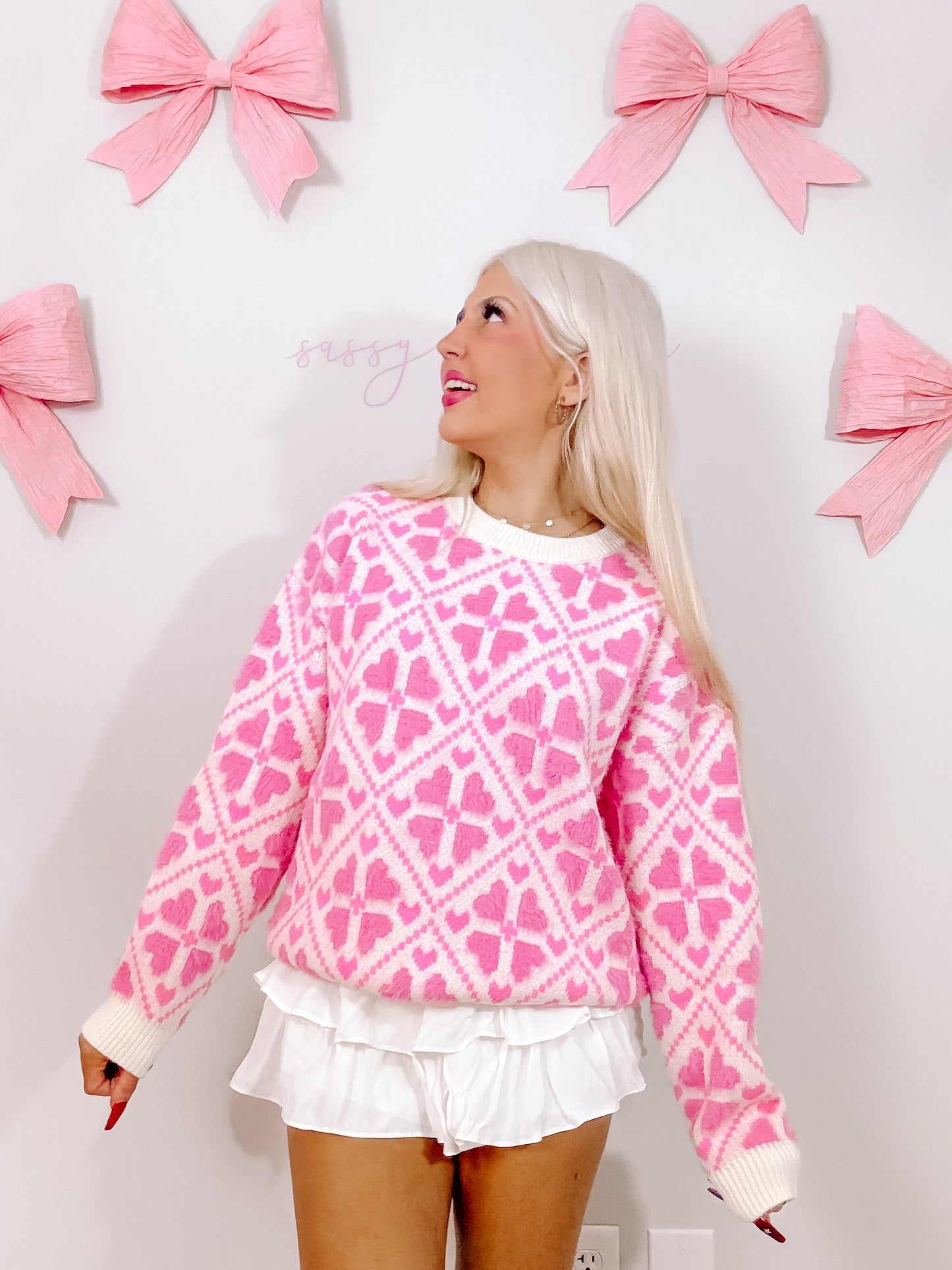 Pink fuzzy sweater with heart pattern, modeled against a wall with pink bows.