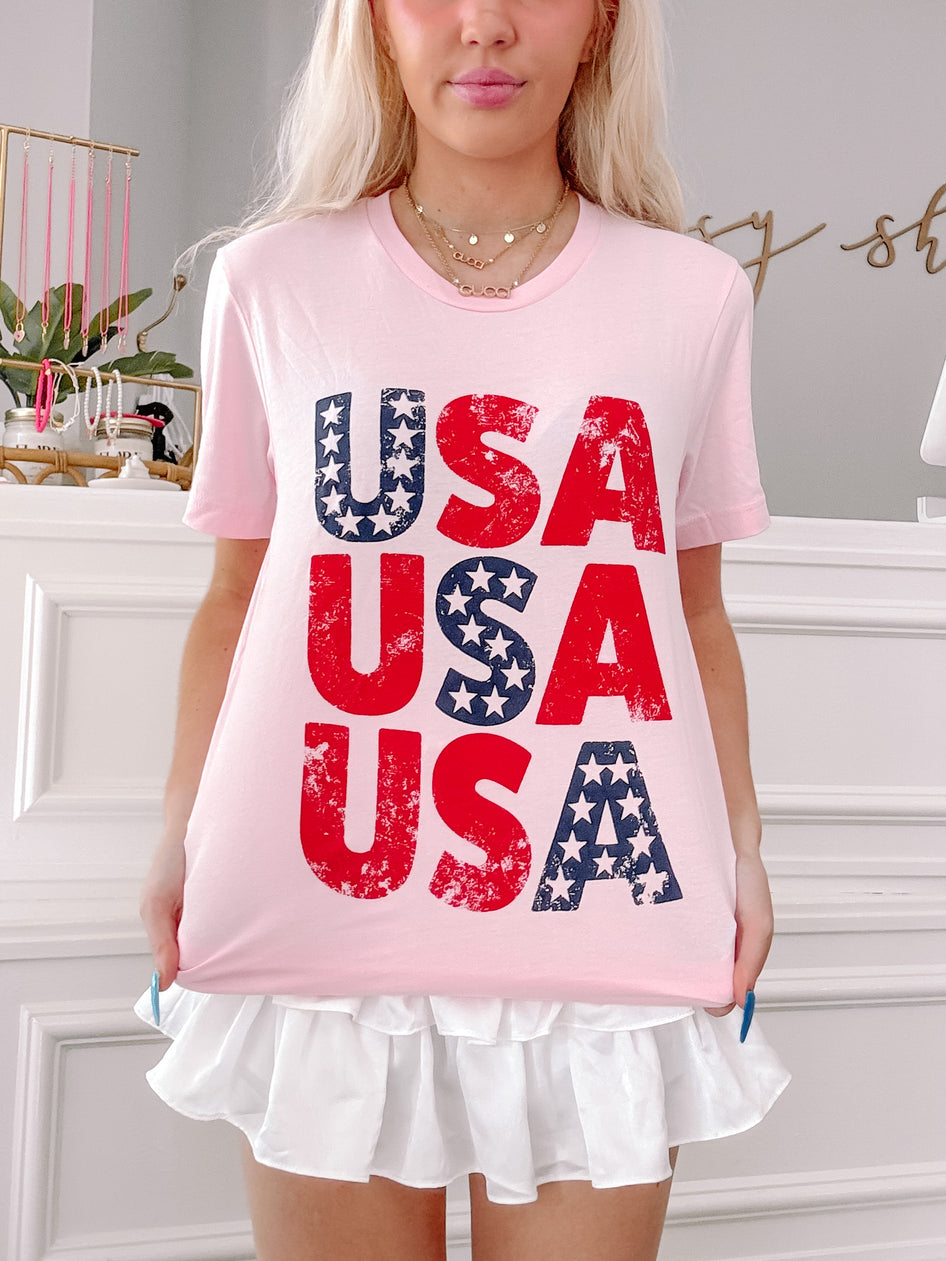 Pink tee featuring a patriotic "USA" graphic with distressed red lettering and navy stars.