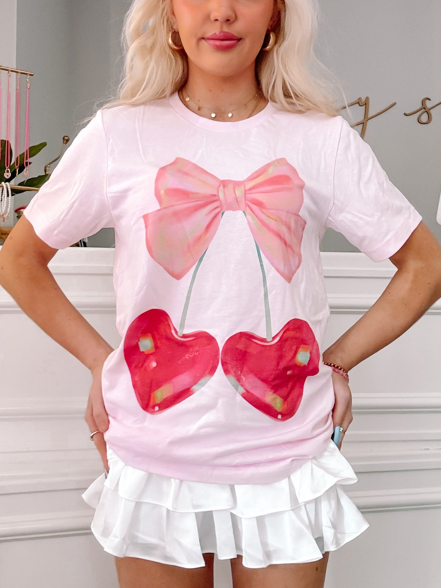 Pink t-shirt featuring a whimsical cherry and bow graphic; model wears white ruffled skirt.
