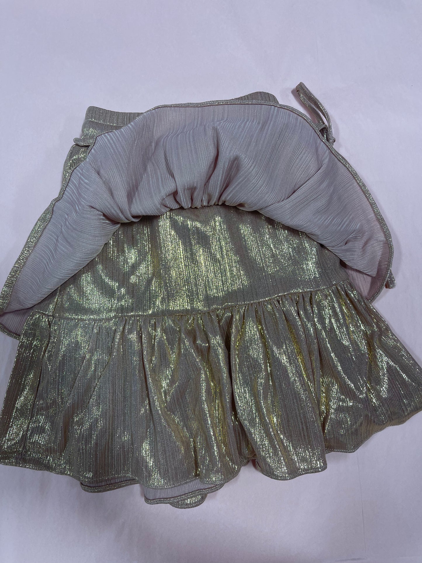 Gold shimmering mini skirt with ruffled hem, shown inside out on a pale pink background.