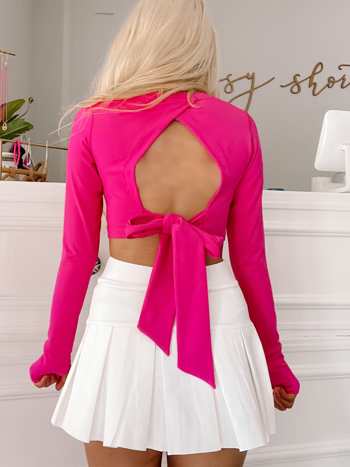 Hot pink long-sleeve crop top with open back and bow tie detail, paired with a white pleated mini skirt.