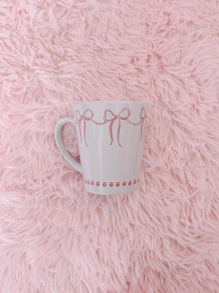 White ceramic mug with pink ribbon design, resting on a fluffy pink surface.