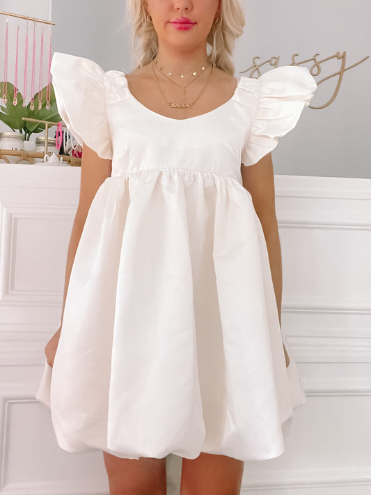 Ivory mini dress with puffed sleeves and a bubble hem, shown on a model against a white wall.