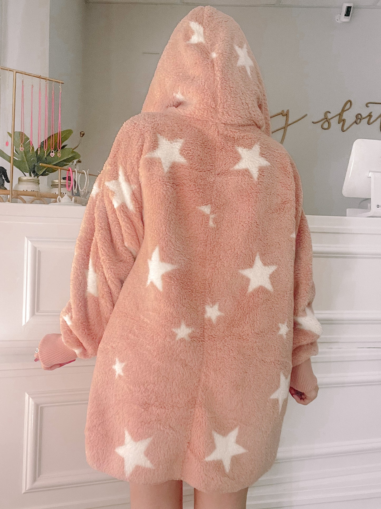 Pink fuzzy hooded jacket adorned with white stars; cozy sweater shown from behind in a shop setting.