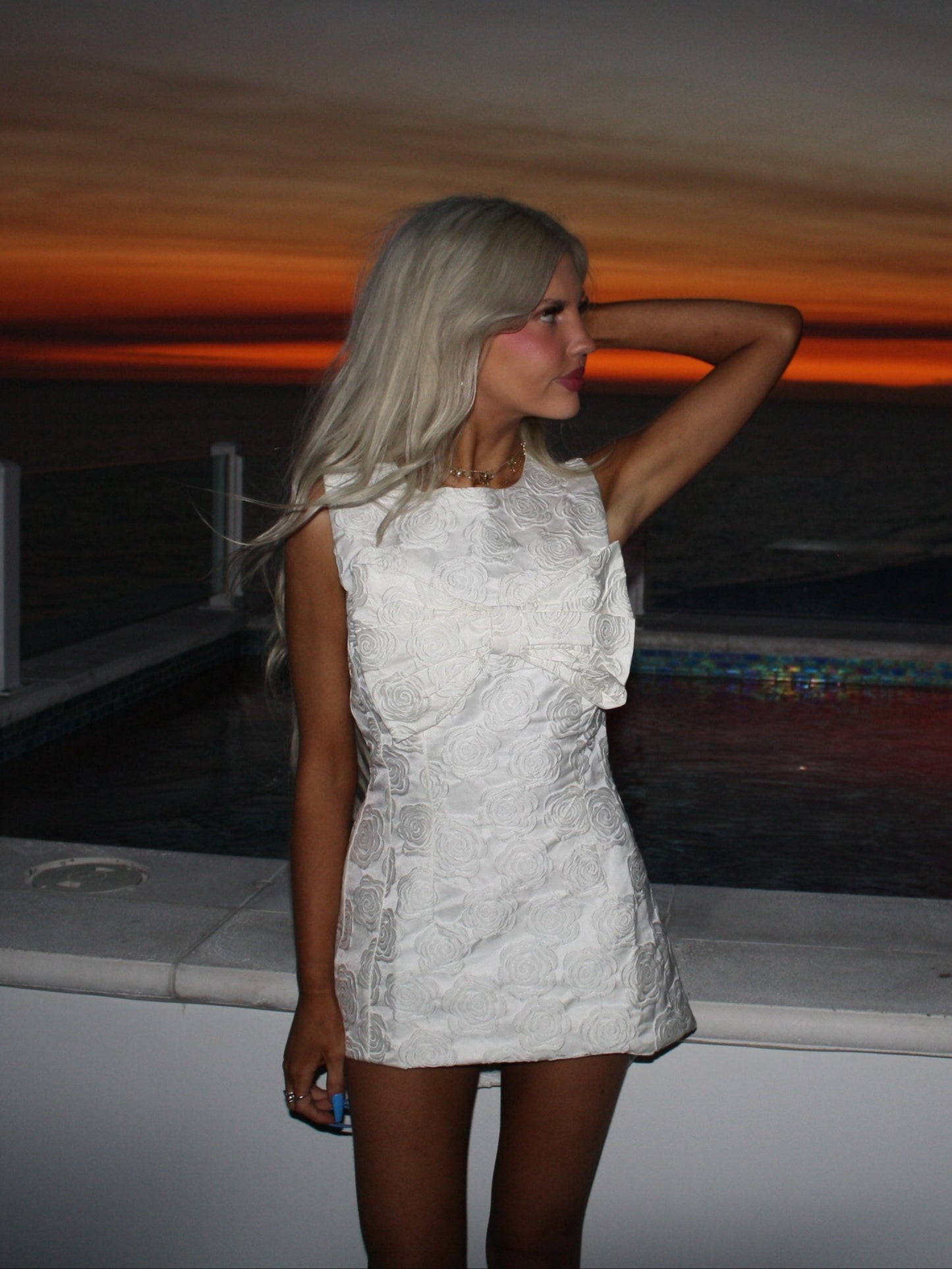 White rose-patterned mini dress, worn by a model at sunset by a pool.