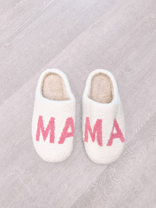 White fluffy slippers with pink "Mama" lettering, shown on a wood floor.