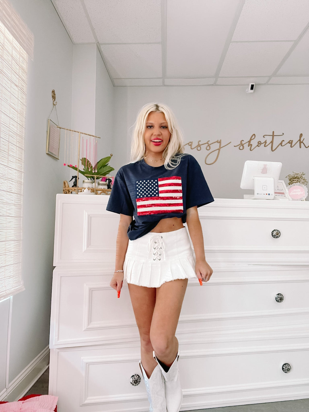 Blue Born in the USA Tee by Sassy Shortcake with red white and blue sequin flag Woman in white skirt and dark tee featuring an American flag graphic, standing in front of white dresser.