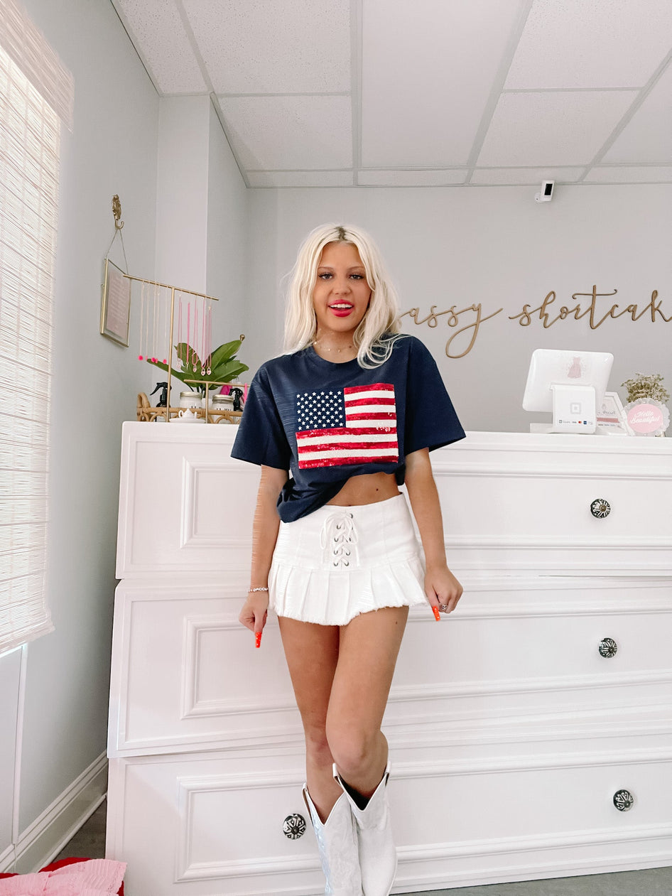 Woman in white pleated mini skirt and blue USA flag graphic tee, posing in a bright retail space.