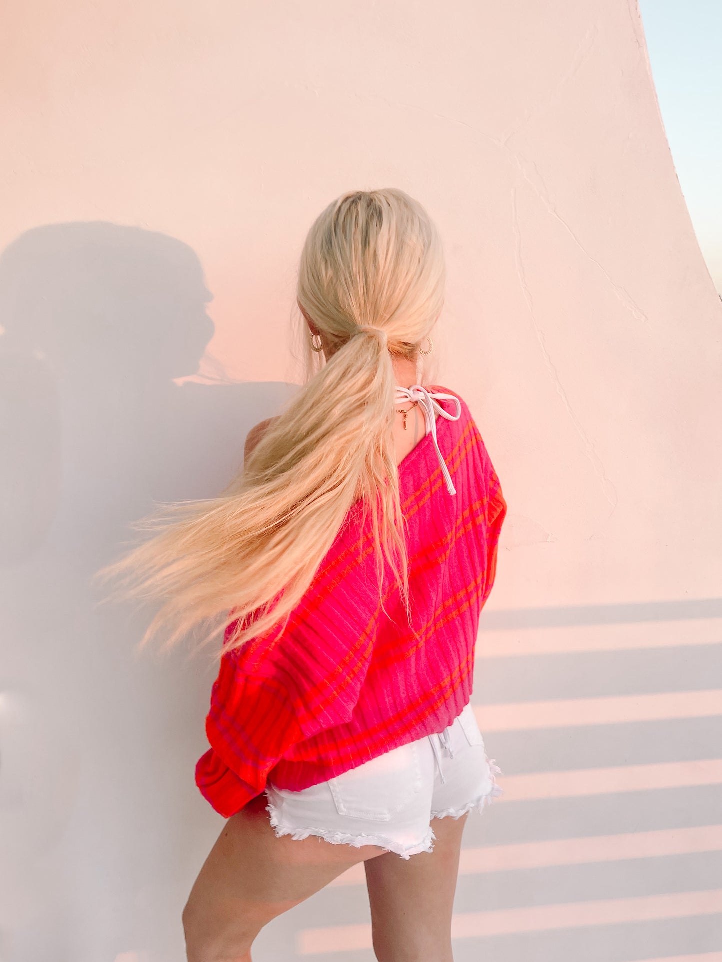 Pink and orange striped knit sweater; back view, worn with white denim shorts, against a light pink wall.