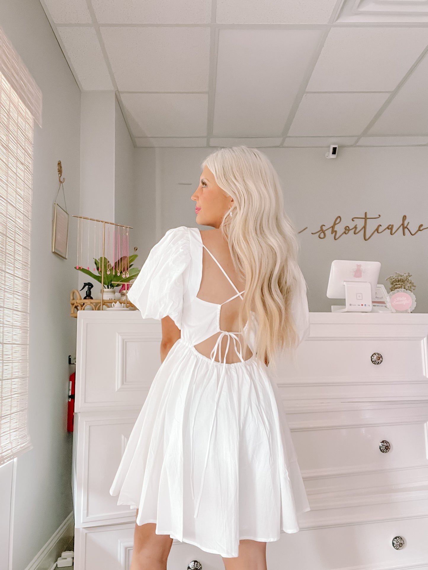 White eyelet mini dress with puff sleeves and open back, shown in a bright interior.