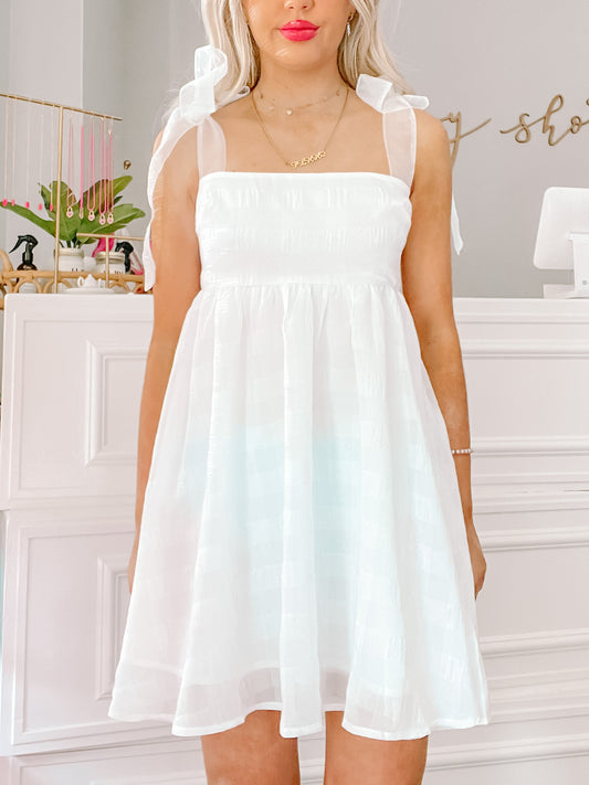 White eyelet mini dress with sheer straps and bow detailing, shown on a model.