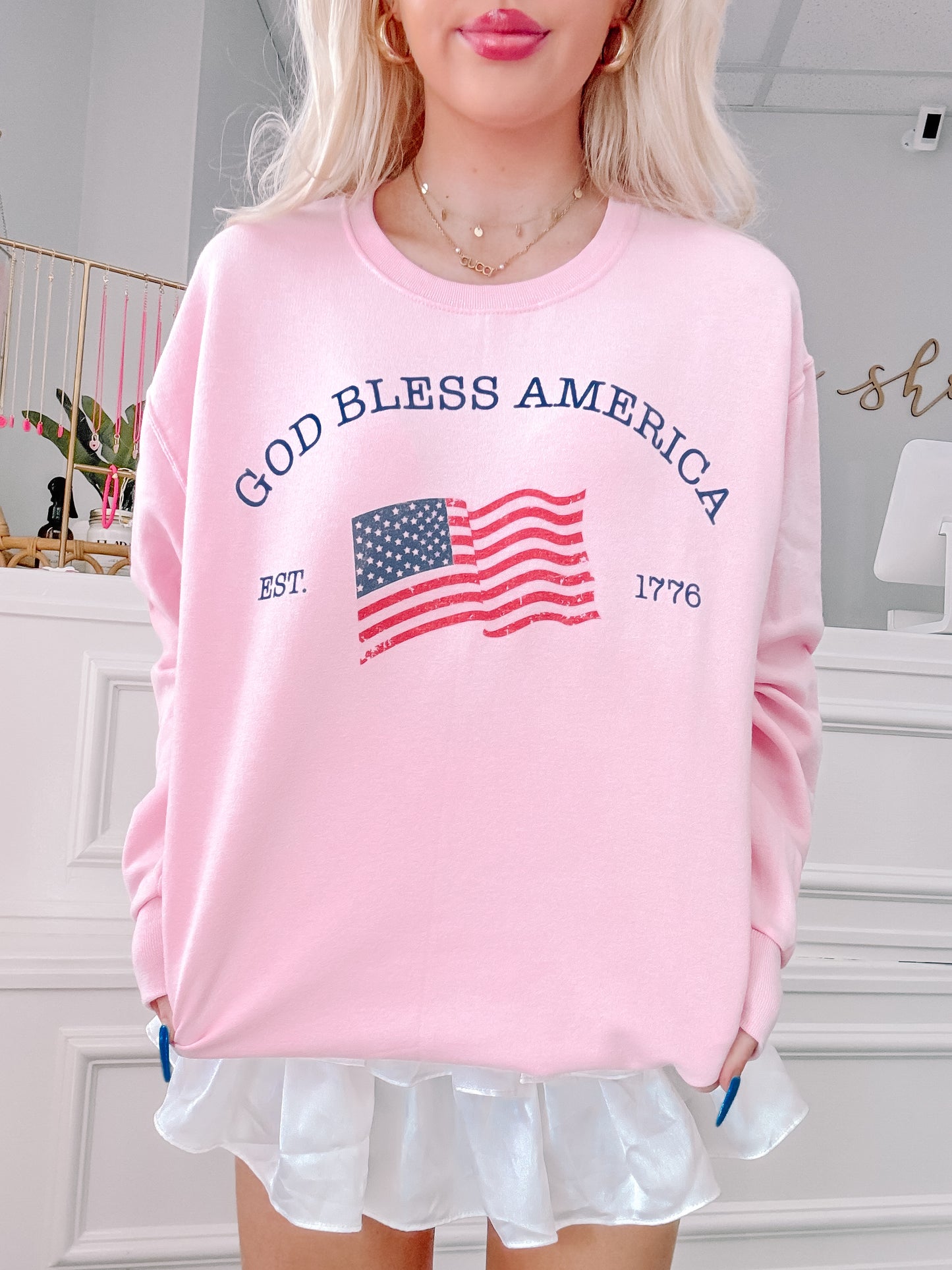 Pink crewneck sweatshirt with an American flag graphic and "God Bless America, Est. 1776" text.