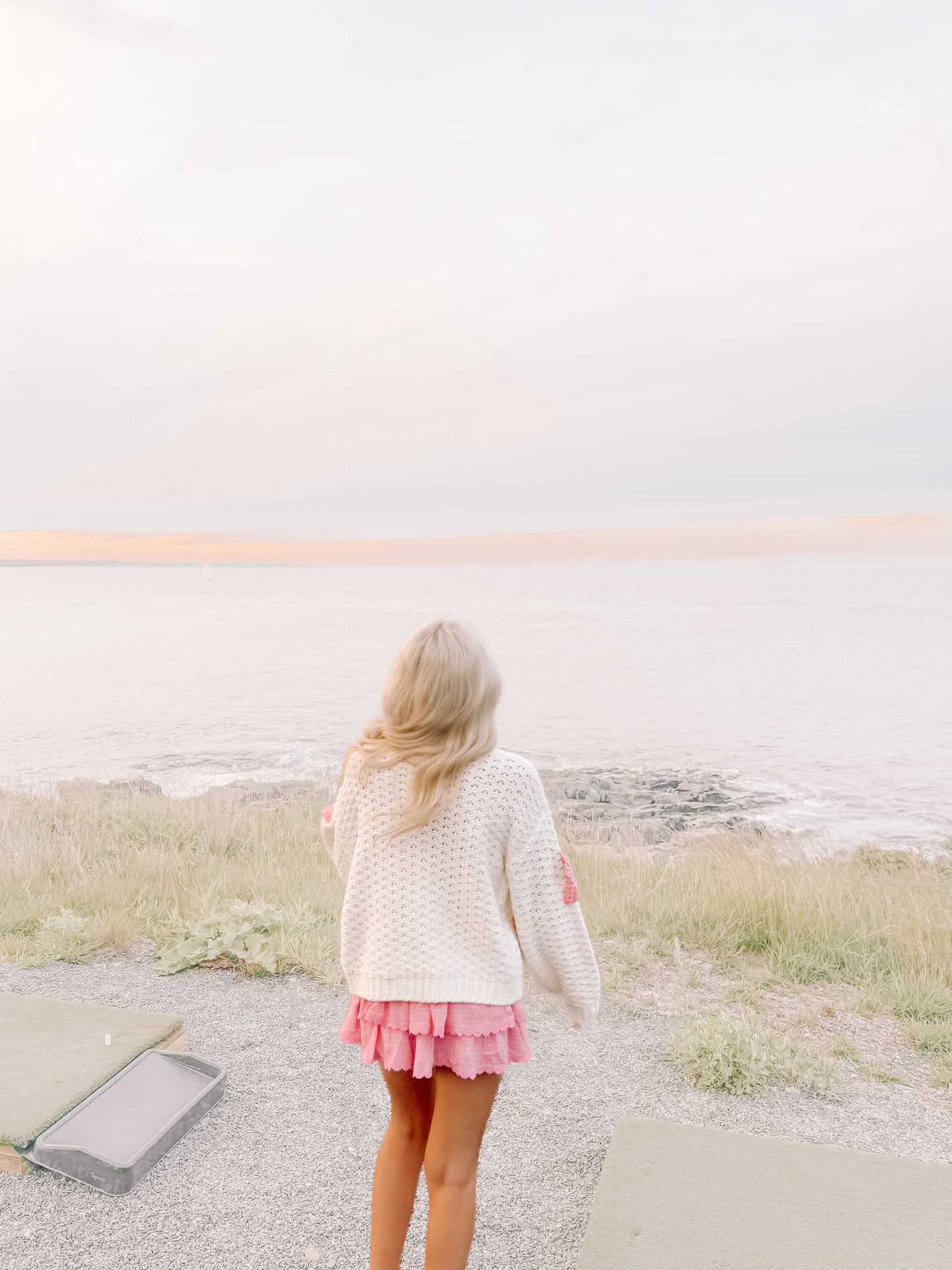Woman in cream knit cardigan and pink skirt overlooking ocean at sunset.