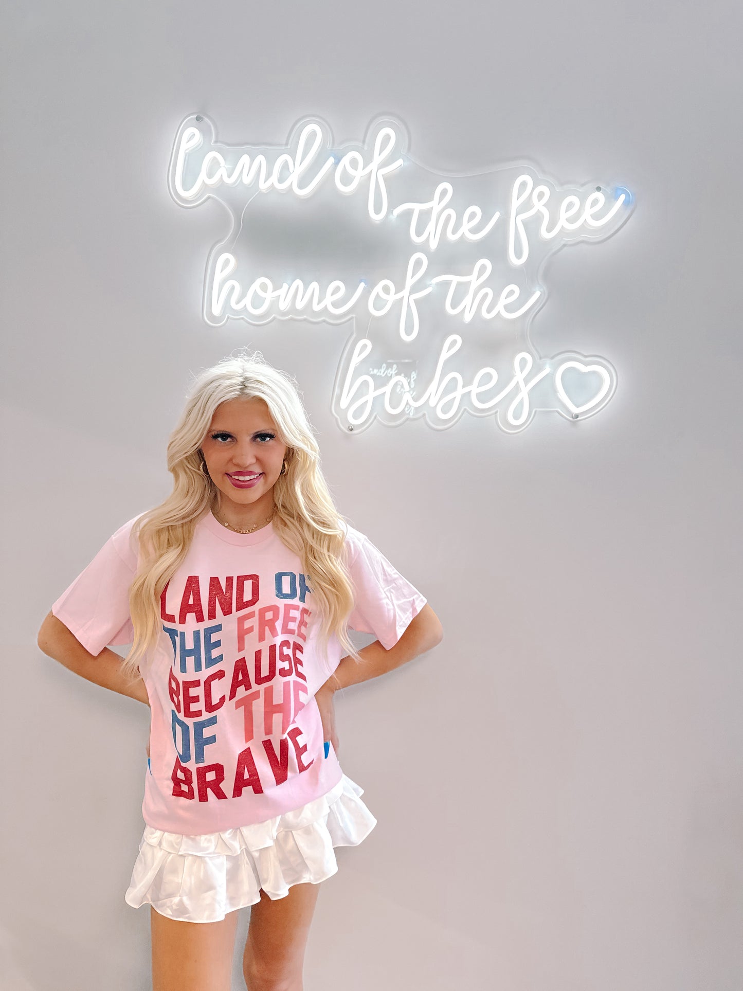 Pink patriotic tee shirt styled with white pleated mini skirt, shown against a wall with a neon sign.
