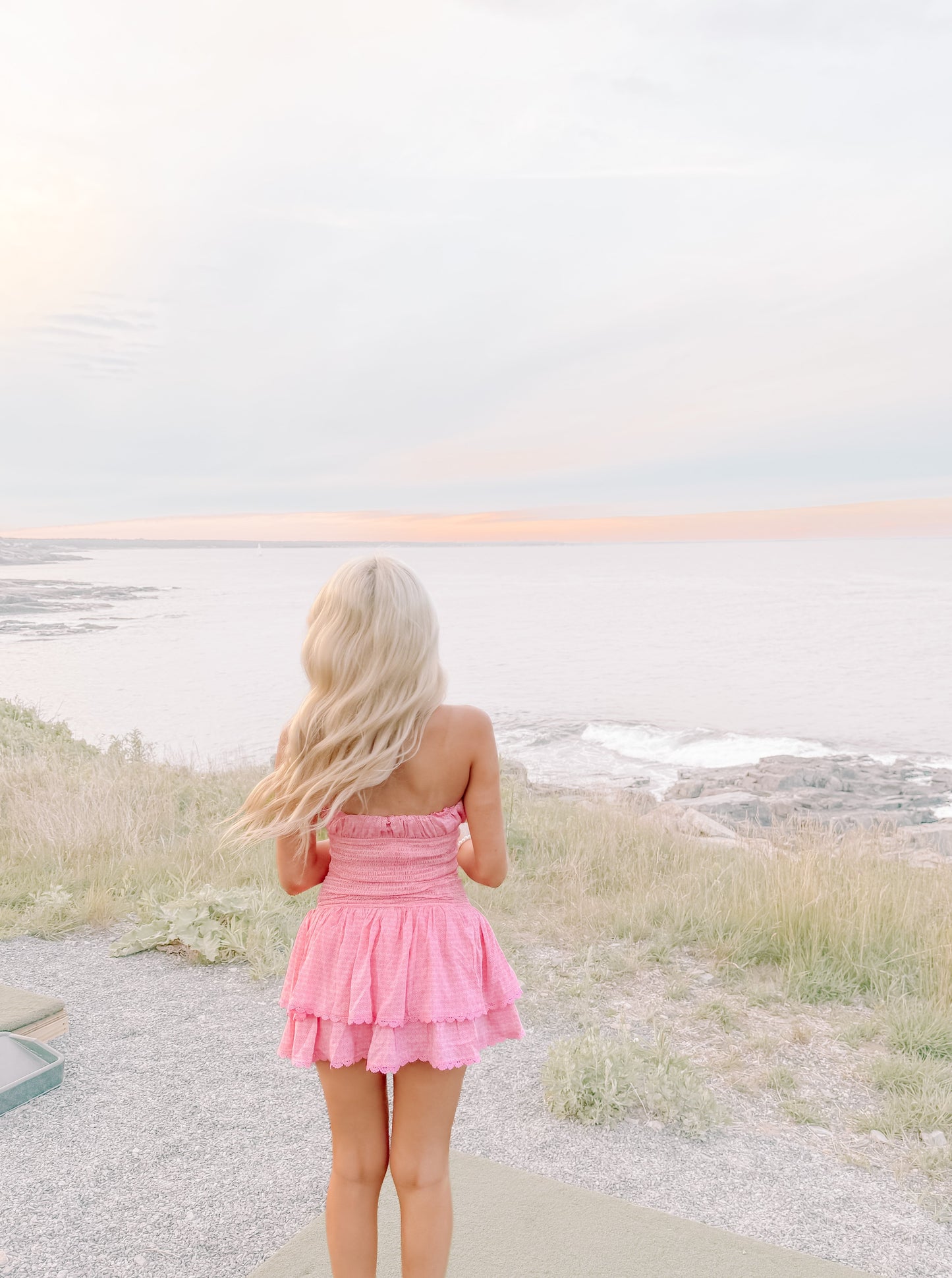 Pink tiered mini dress viewed from behind, overlooking a tranquil ocean at sunset.