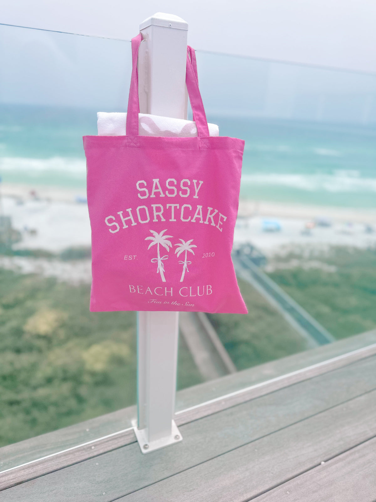 Pink canvas tote bag hanging outdoors, palm tree design, ocean view in background. Sassy Shortcake Beach Club Tote