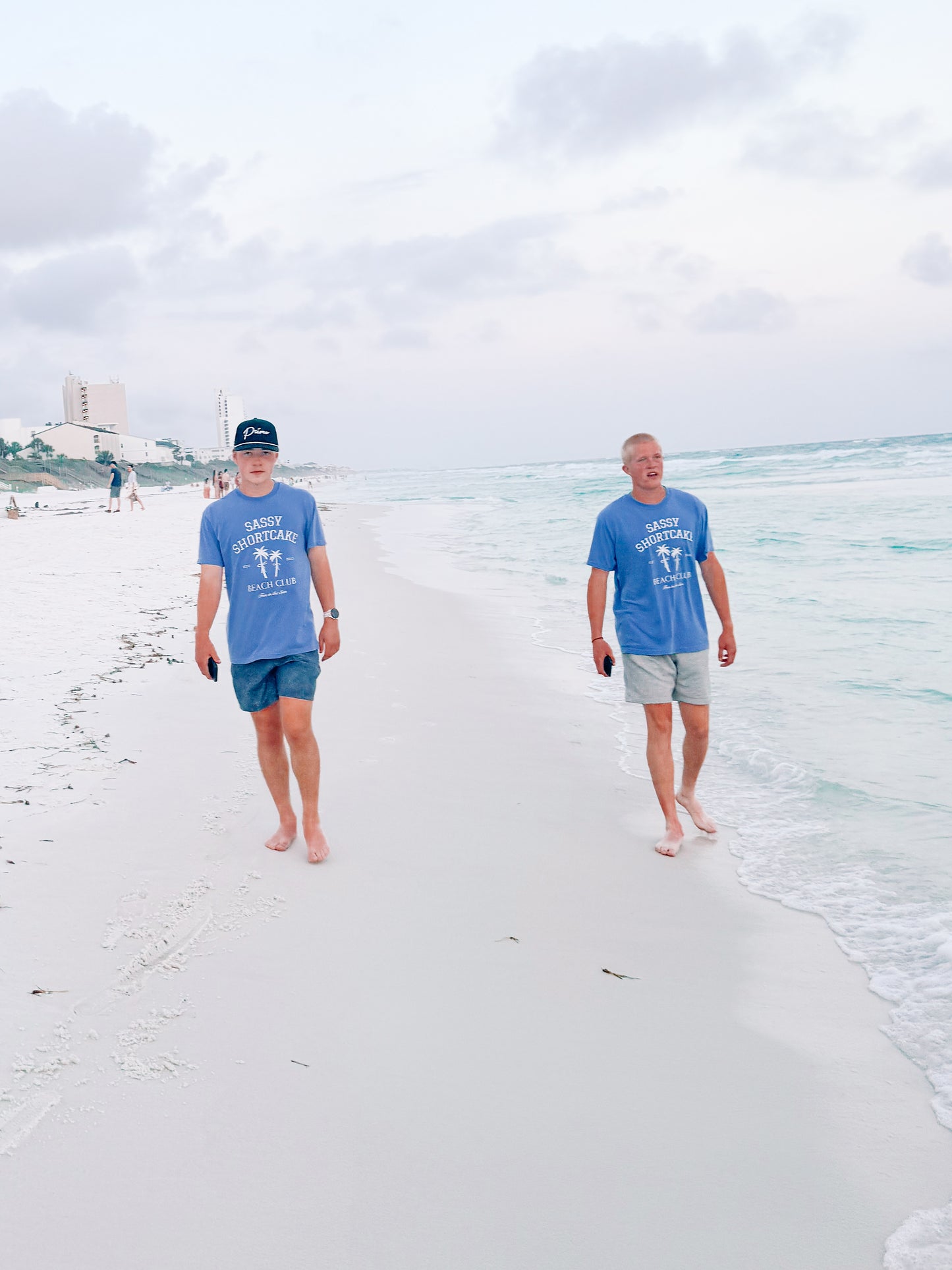Two men stroll along a sandy beach, wearing matching light blue short-sleeved shirts, ocean waves gently lapping at the shore.