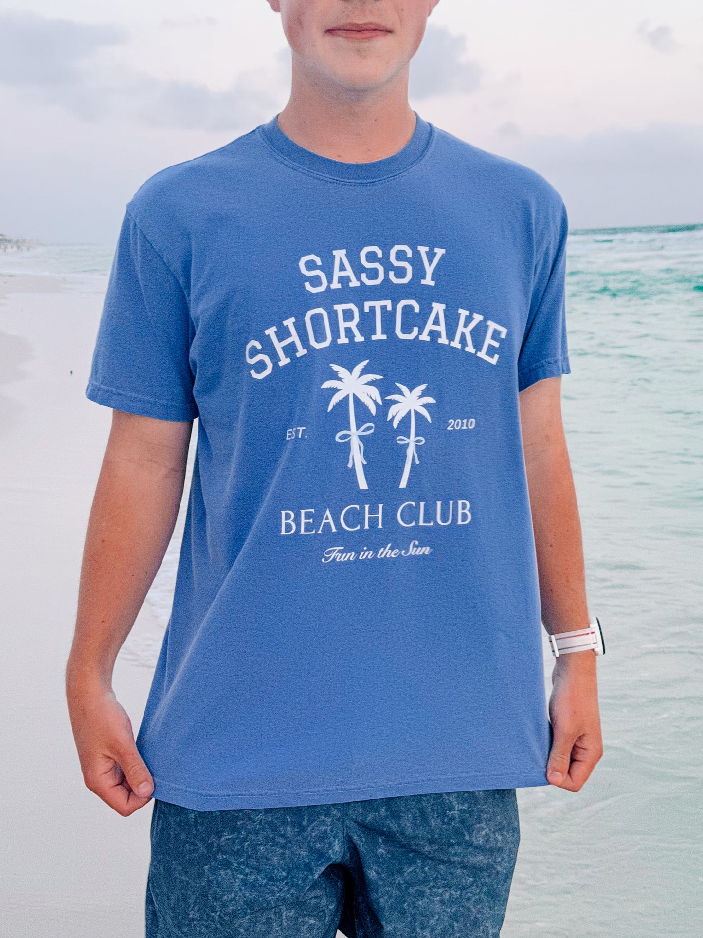 Blue beach-themed tee shirt featuring a palm tree graphic, displayed outdoors near the ocean.
