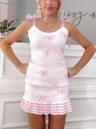 Cream knit mini dress adorned with pink bows, featuring gingham straps and a pink and white striped hem.