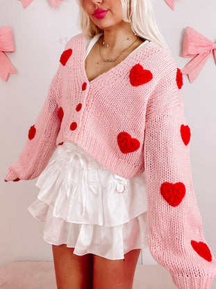 Pink knit cardigan adorned with red felt hearts, modeled with a white ruffled skirt.
