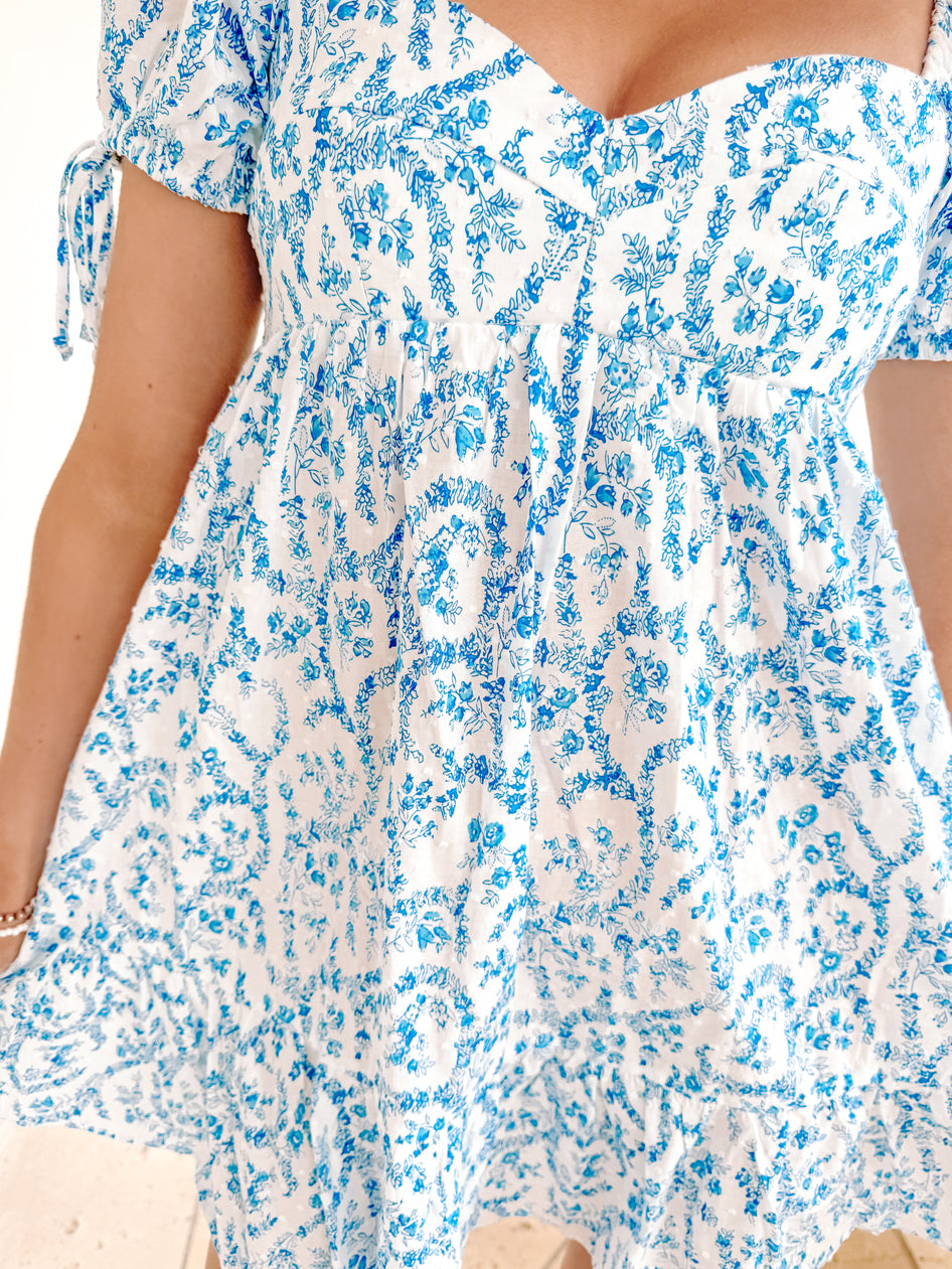 White eyelet mini dress with blue floral print, puff sleeves and tie detail.