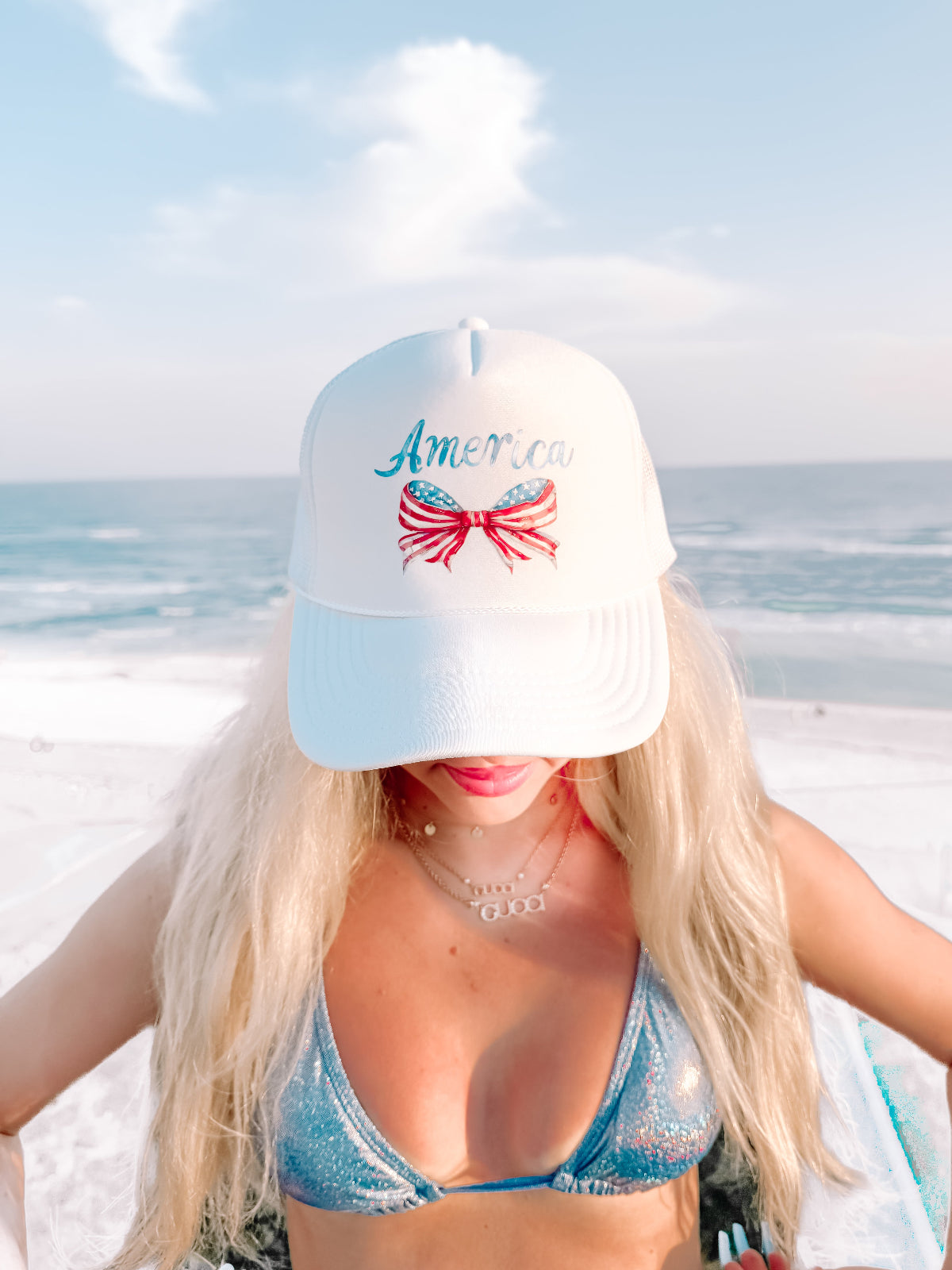 White trucker hat with an American flag bow and "America" script from Sassy Shortcake, beach backdrop