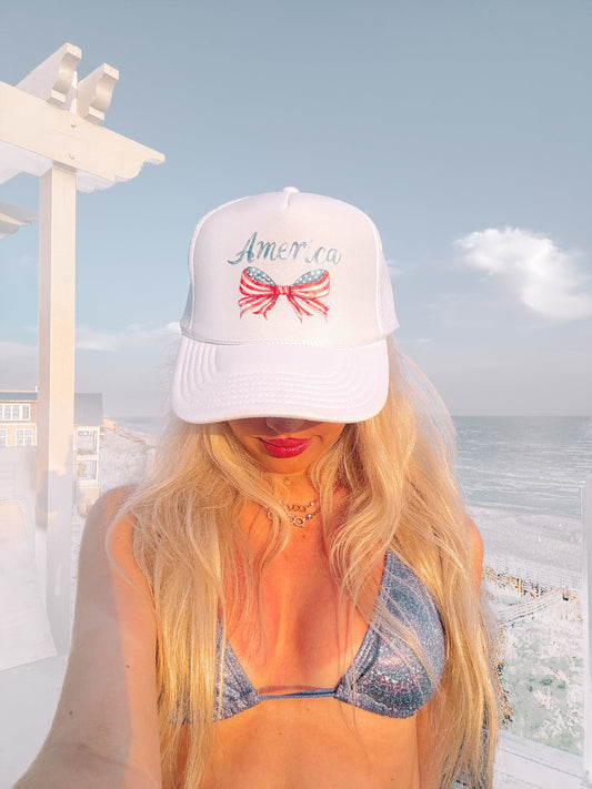 White trucker hat from Sassy Shortcake with a patriotic bow and "America" script, worn outdoors near the ocean