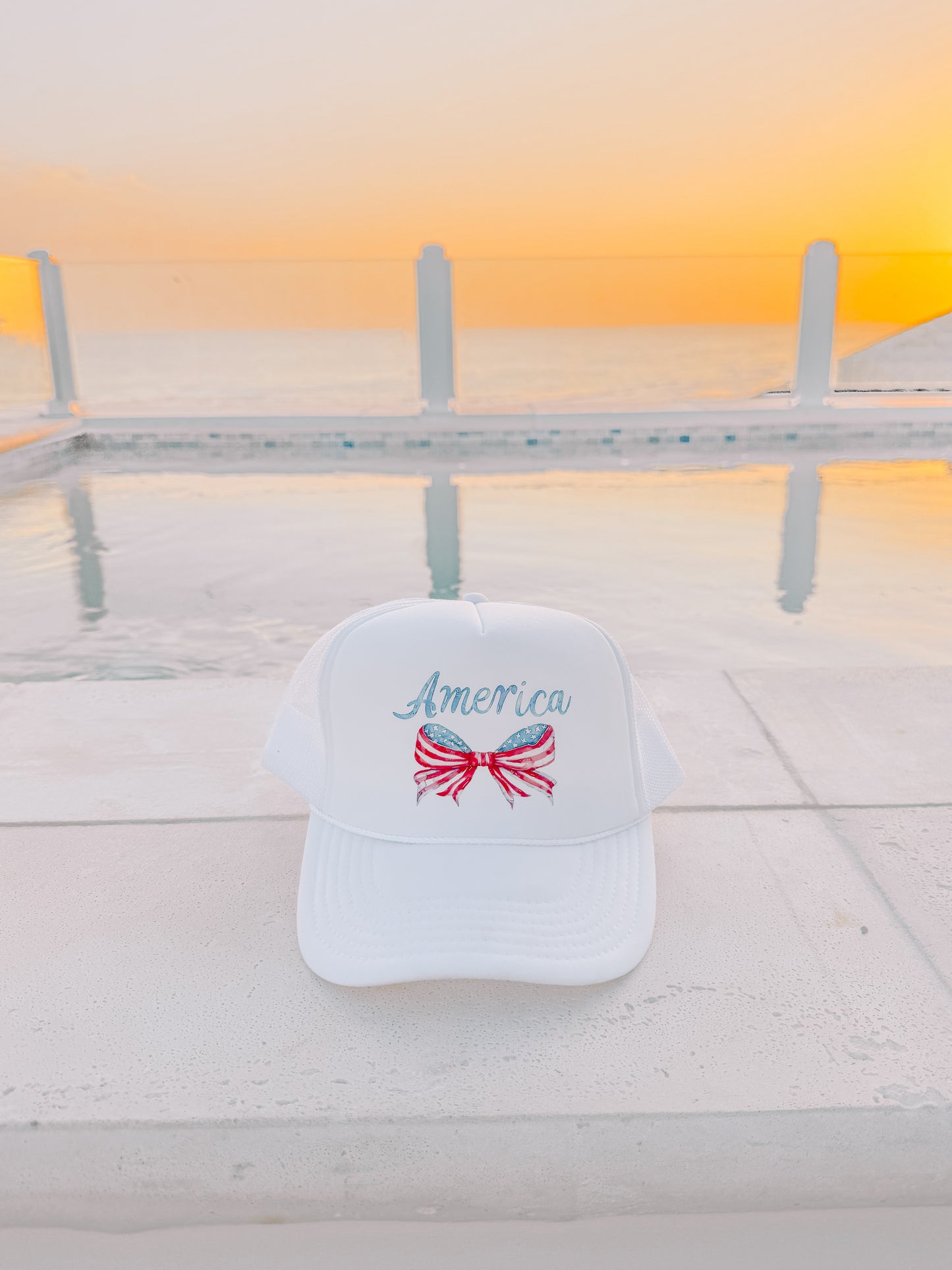 White trucker hat with patriotic bow and text, by a pool at sunset.