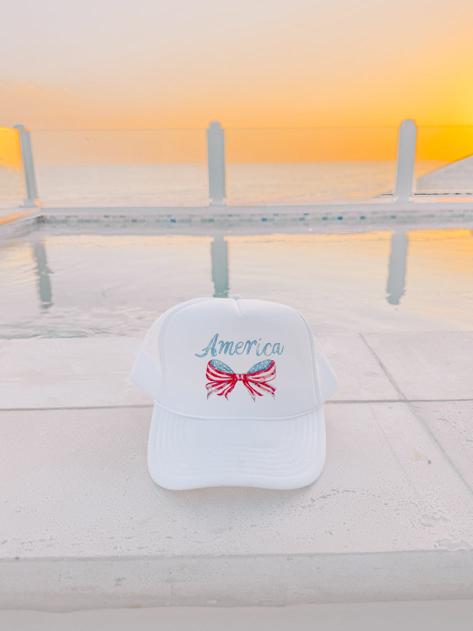 White trucker hat with patriotic bow and text, by a pool at sunset.