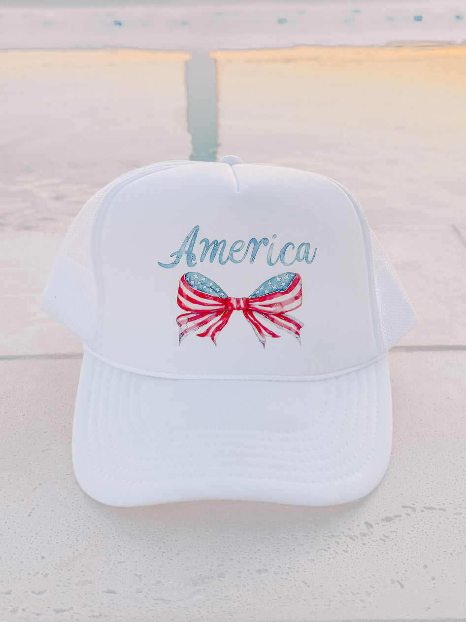 White trucker hat with patriotic bow and "America" design, shown outdoors near water.