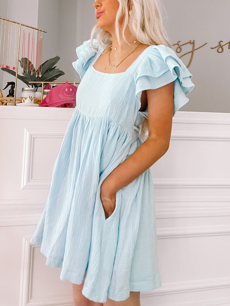 Light blue mini dress with ruffled sleeves, worn against a white paneled wall.