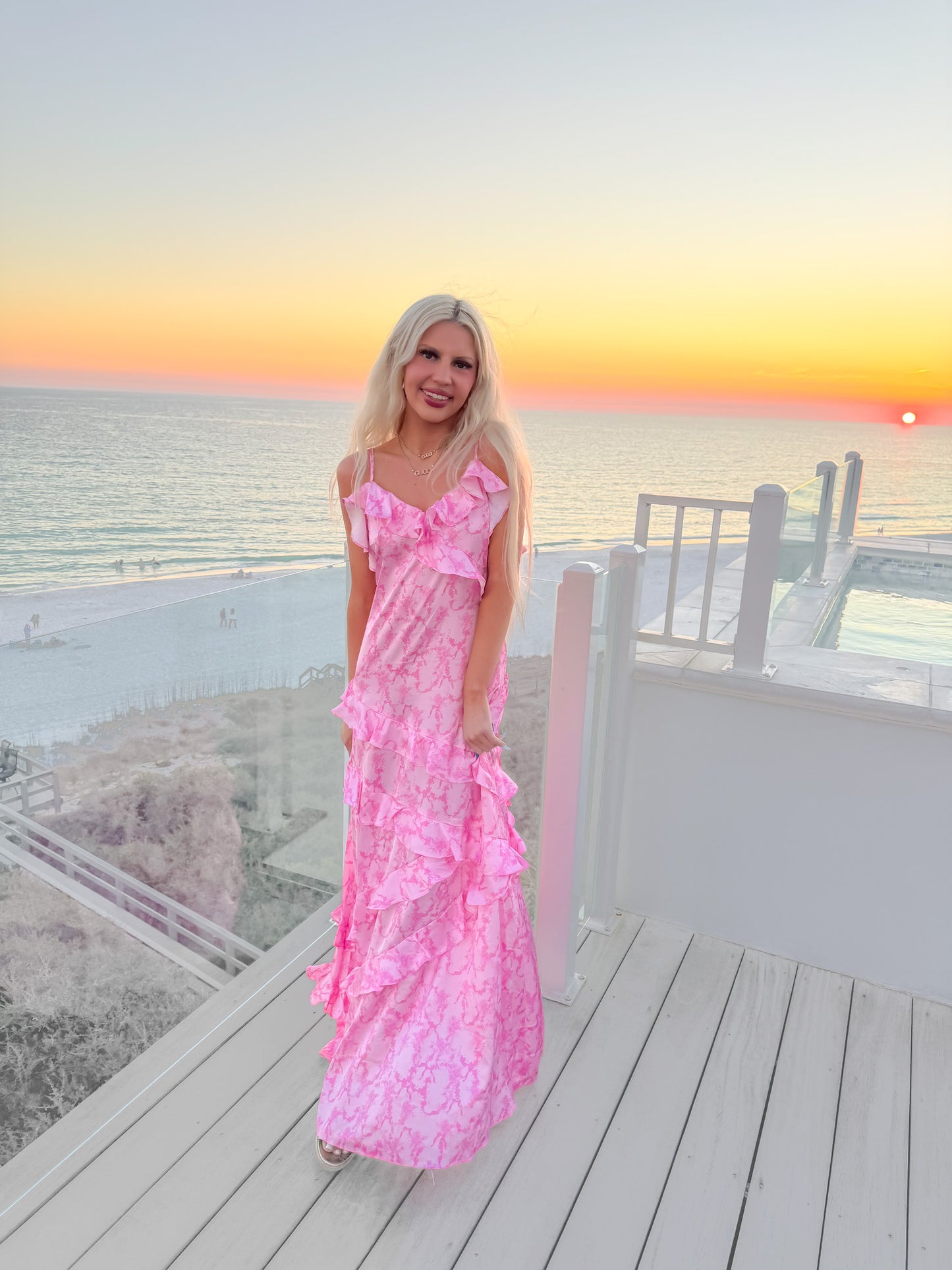 Pink tiered maxi dress with ruffled detailing, ocean sunset backdrop.