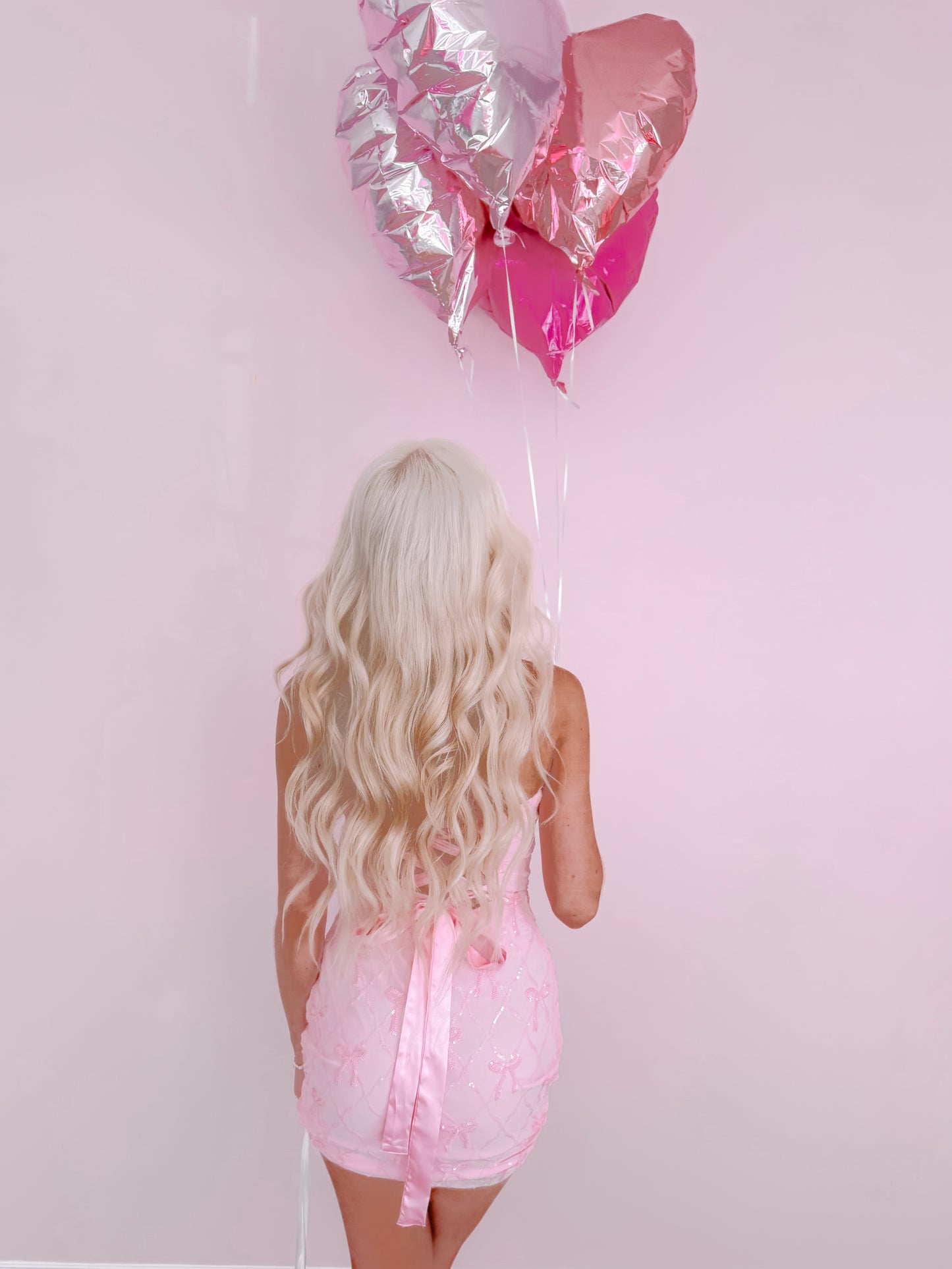 Blond model's back, wearing a pink mini dress against a pink backdrop, holding pink and silver heart-shaped balloons.