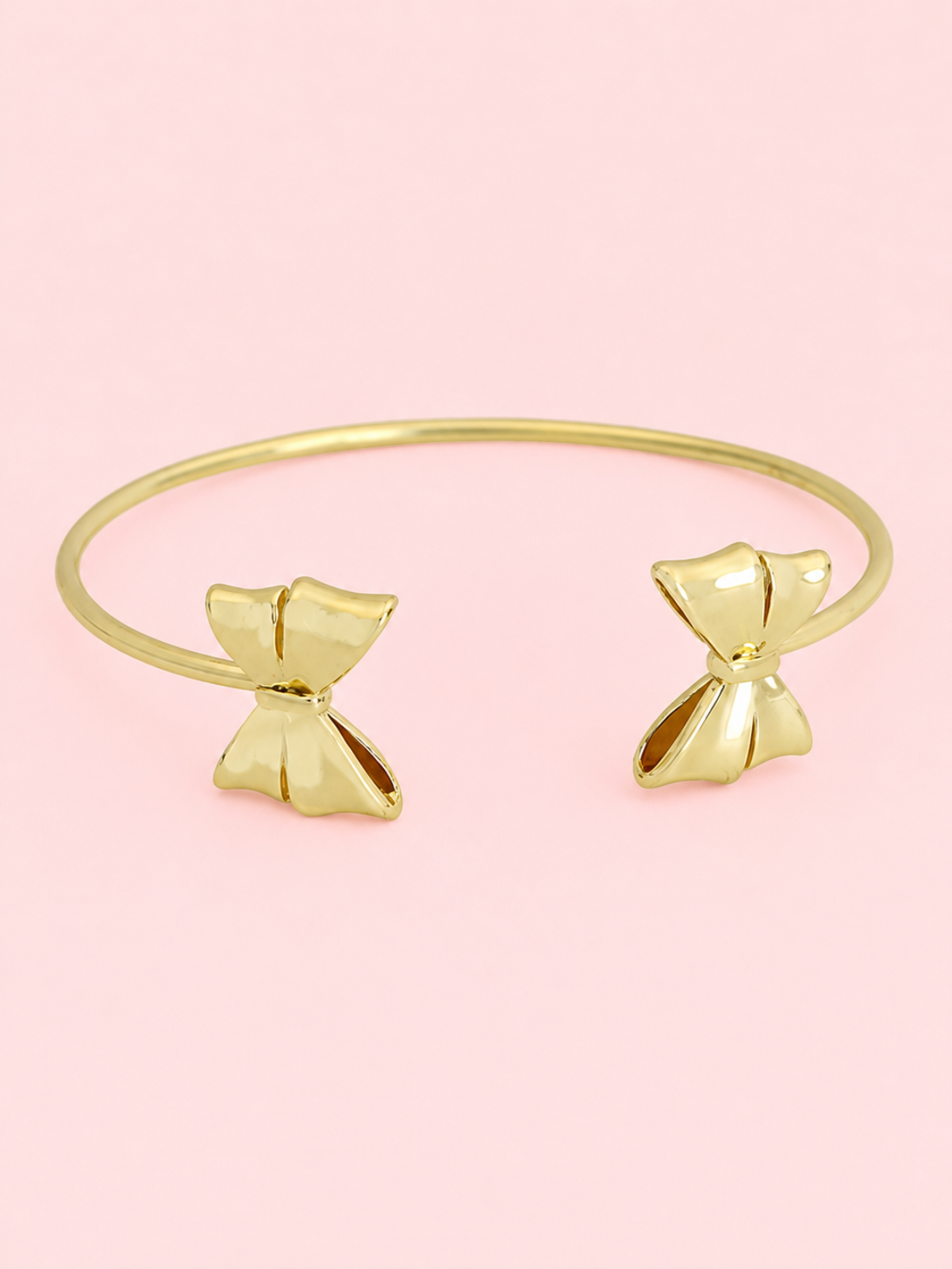 Wrapped In Gold Bow Cuff from Sassy Shortcake: Gold open cuff bracelet with bow details on each end displayed on a light pink background.