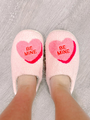 Pink fuzzy slippers with "Be Mine" heart design, shown on a wood floor.