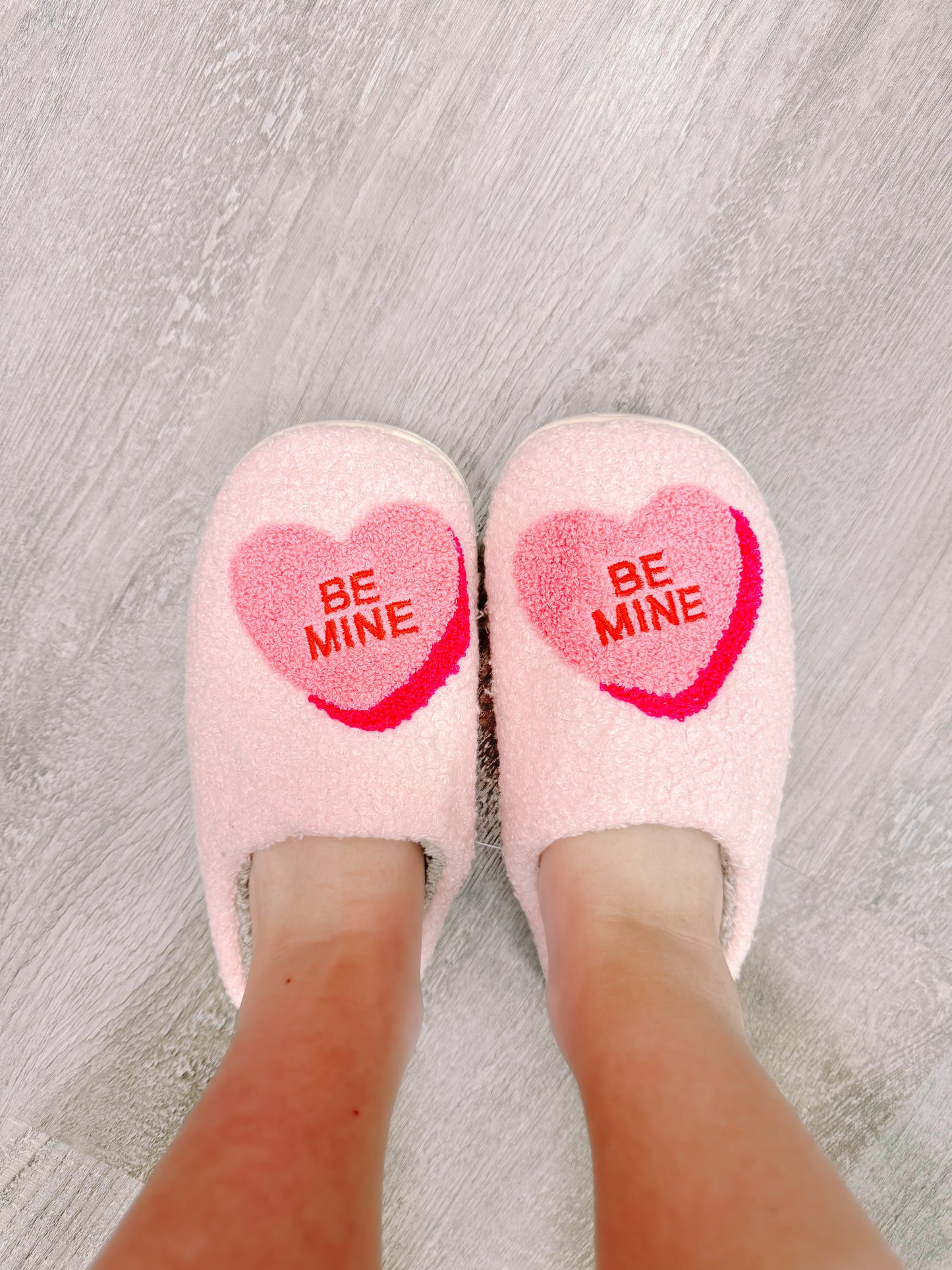 Pink "Be Mine" heart slippers on a wood floor.