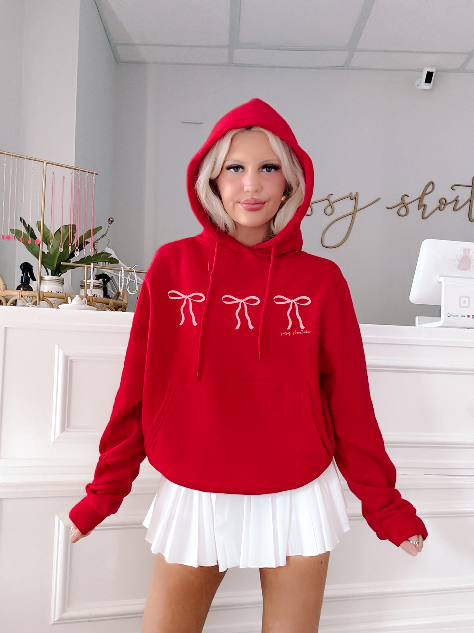 Red hoodie featuring delicate pink bows, modeled indoors against a white backdrop.