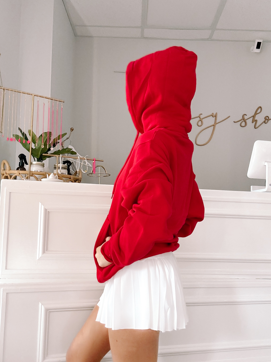 Red hooded sweatshirt styled with a white pleated skirt, shown in a boutique setting.