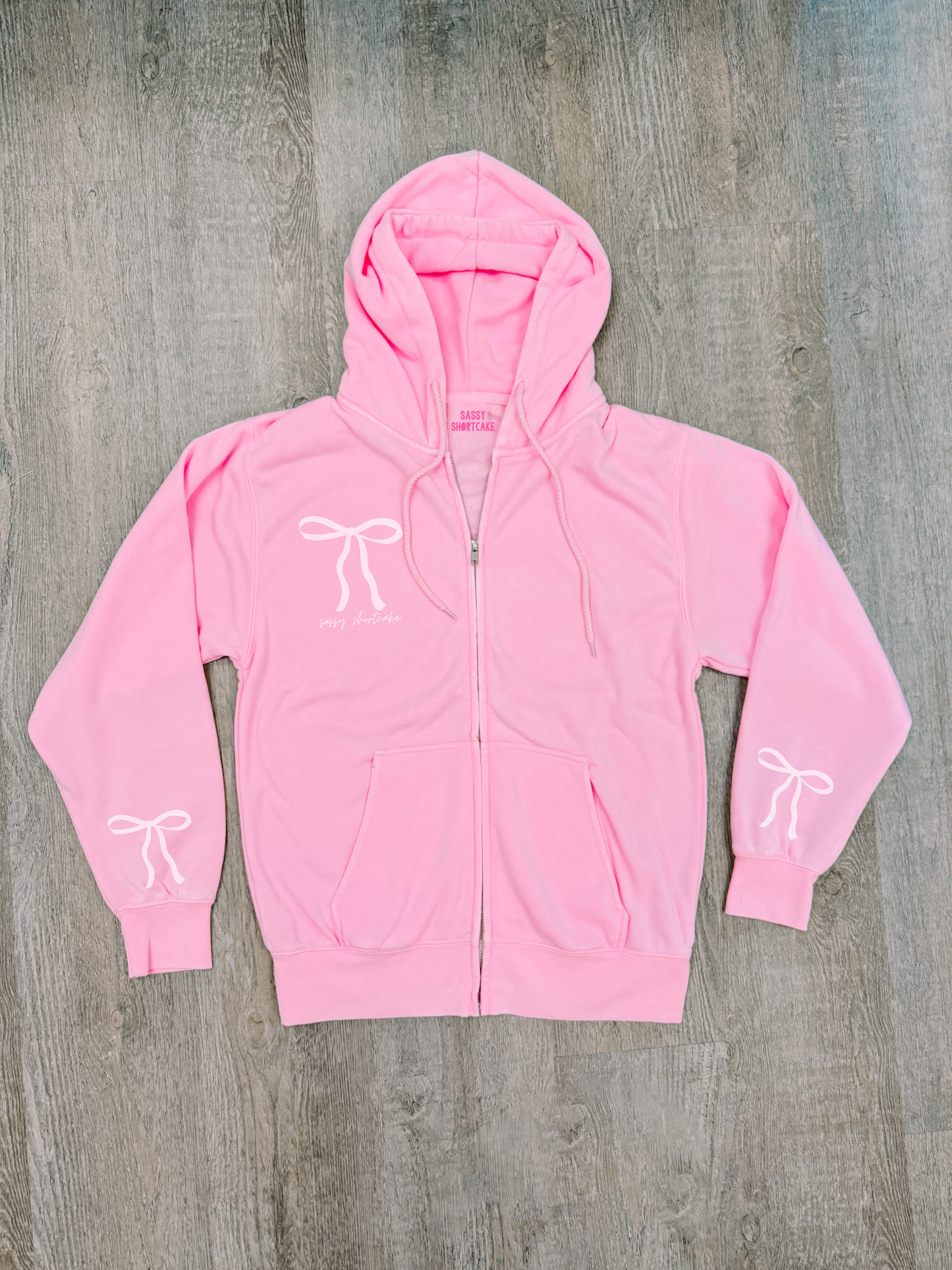 Bubblegum pink zip-up hoodie with bow details, shown flat lay on wood.