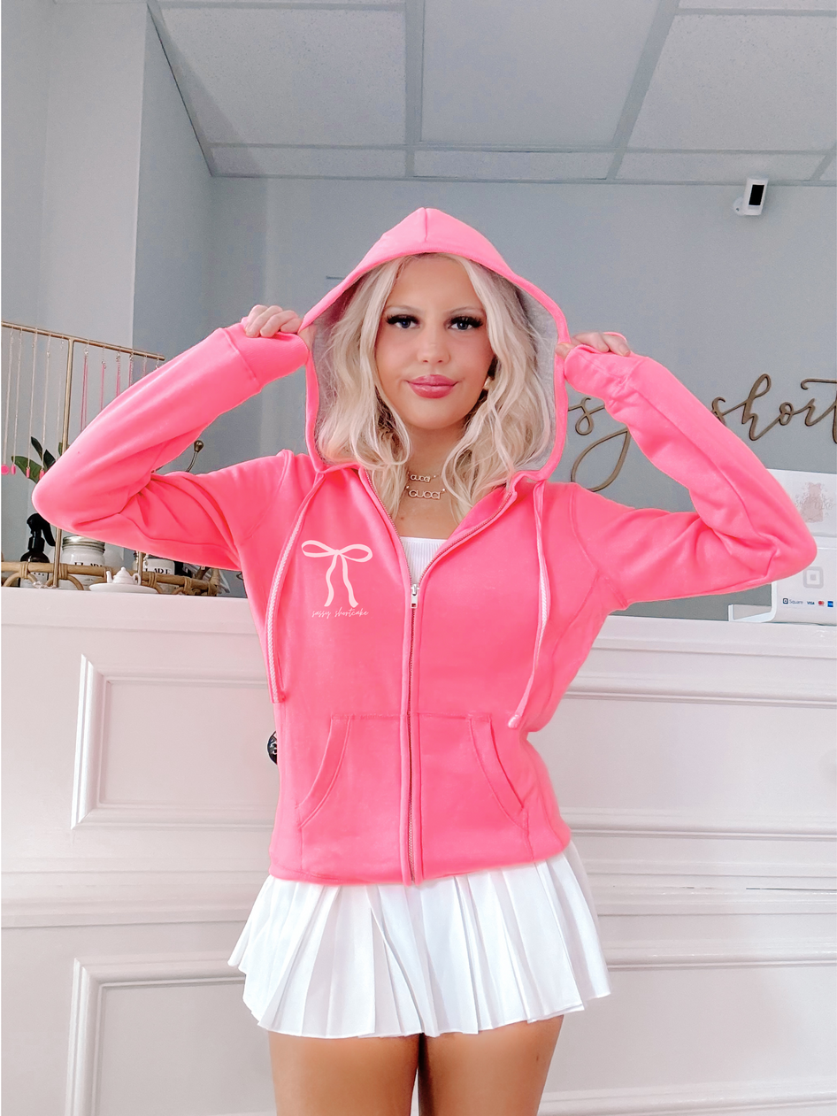 Neon pink zip-up hoodie with bow detail, modeled against a white wall.