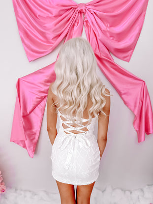 White mini dress with bow details, lace-up back, shown against a pink satin backdrop.