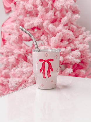 Pink tumbler with a red bow design, featuring a metal straw, set against a blurry pink background.