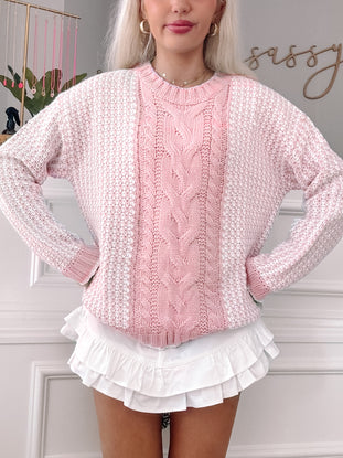 Pink and white cable-knit sweater with textured detailing, styled with a ruffled white skirt.