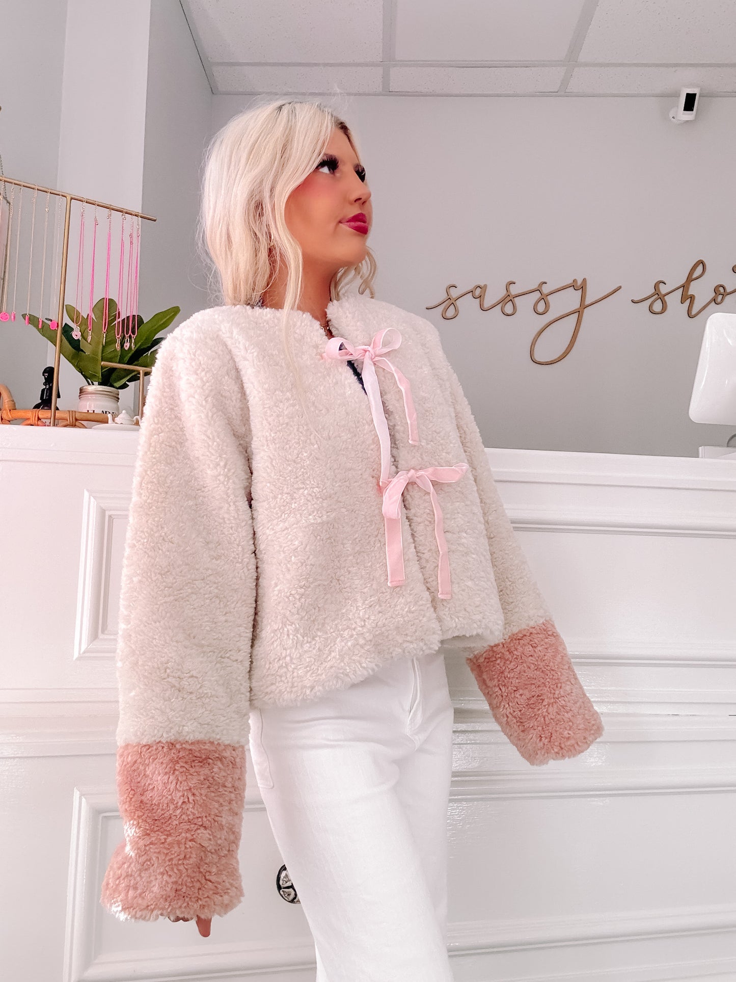 Cream sherpa jacket with pink cuffs and satin ribbon detail, shown on model in boutique setting.