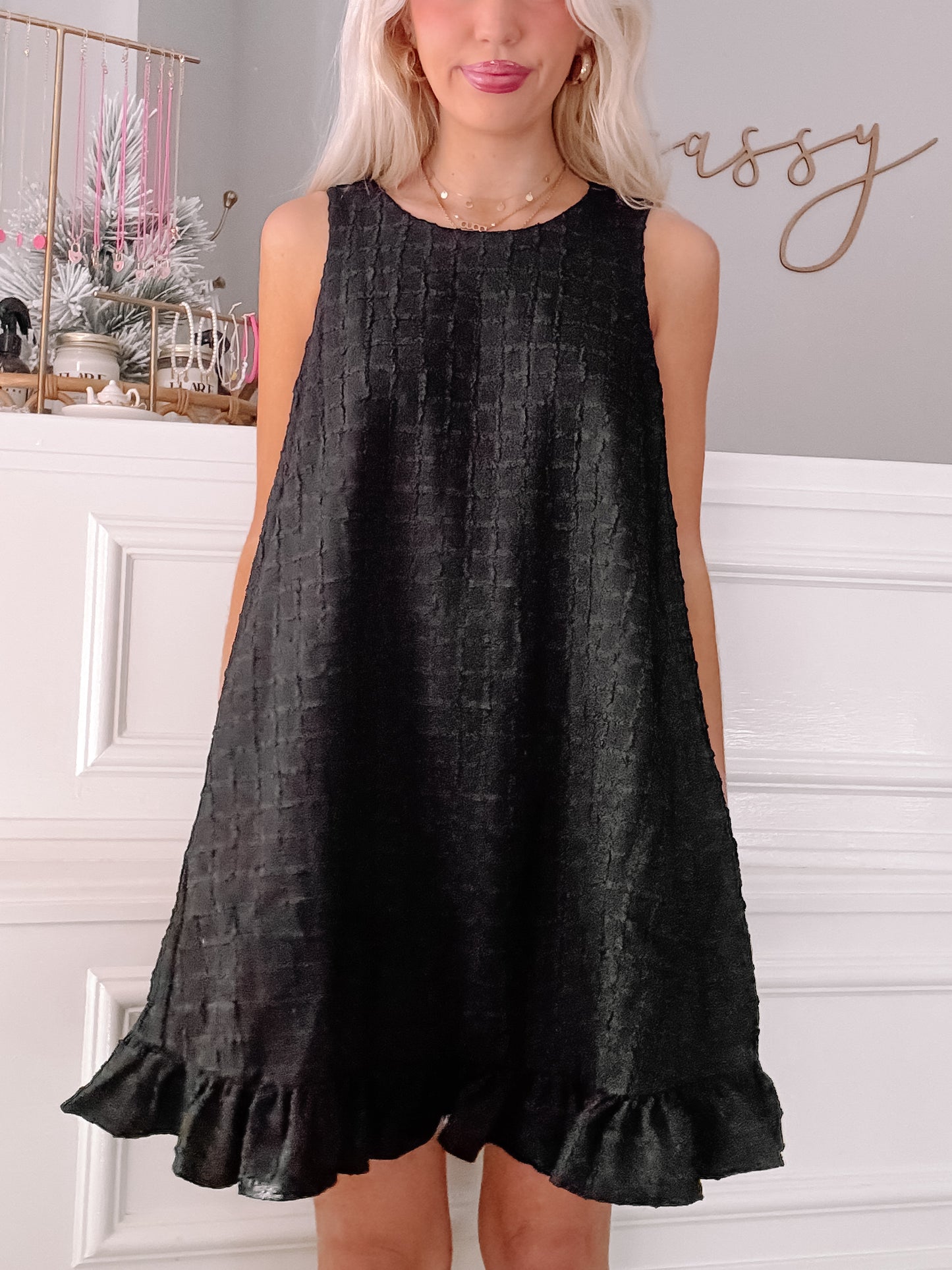 Black sleeveless mini dress with textured fabric and ruffled hem, modeled against a white wall.