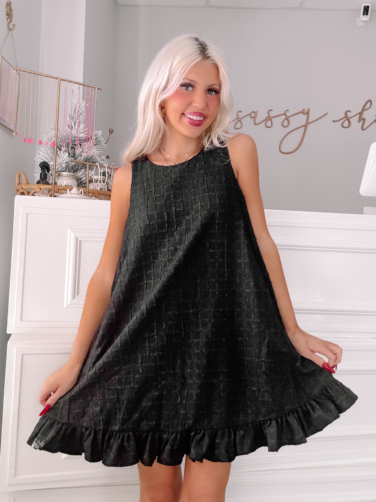 Black sleeveless mini dress with textured fabric and ruffled hem, modeled in a boutique.