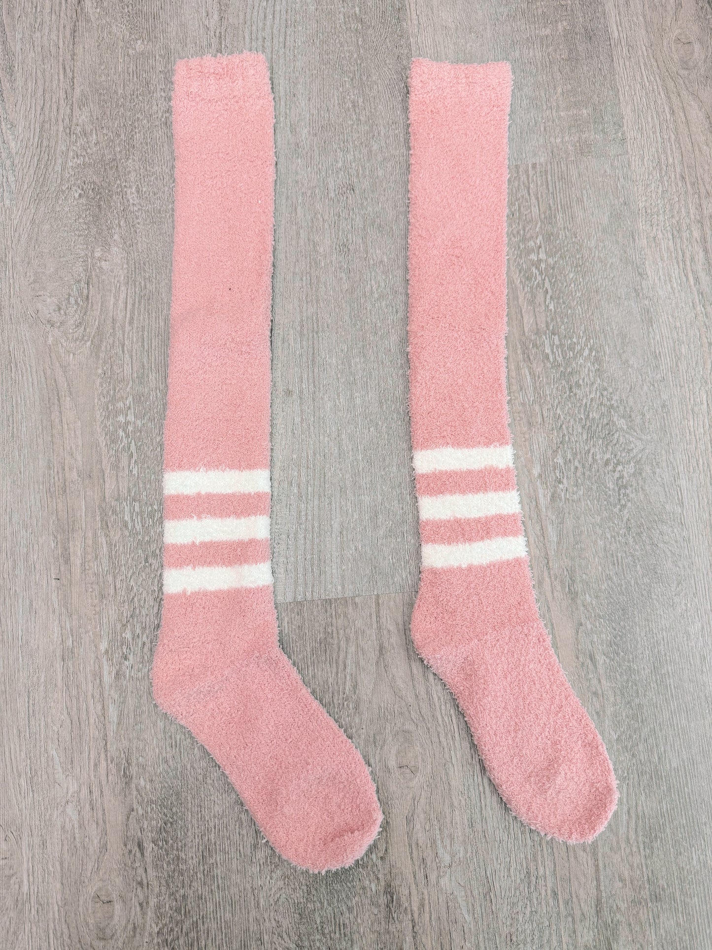 Pair of fluffy pink knee-high socks with white stripes, displayed on wood.