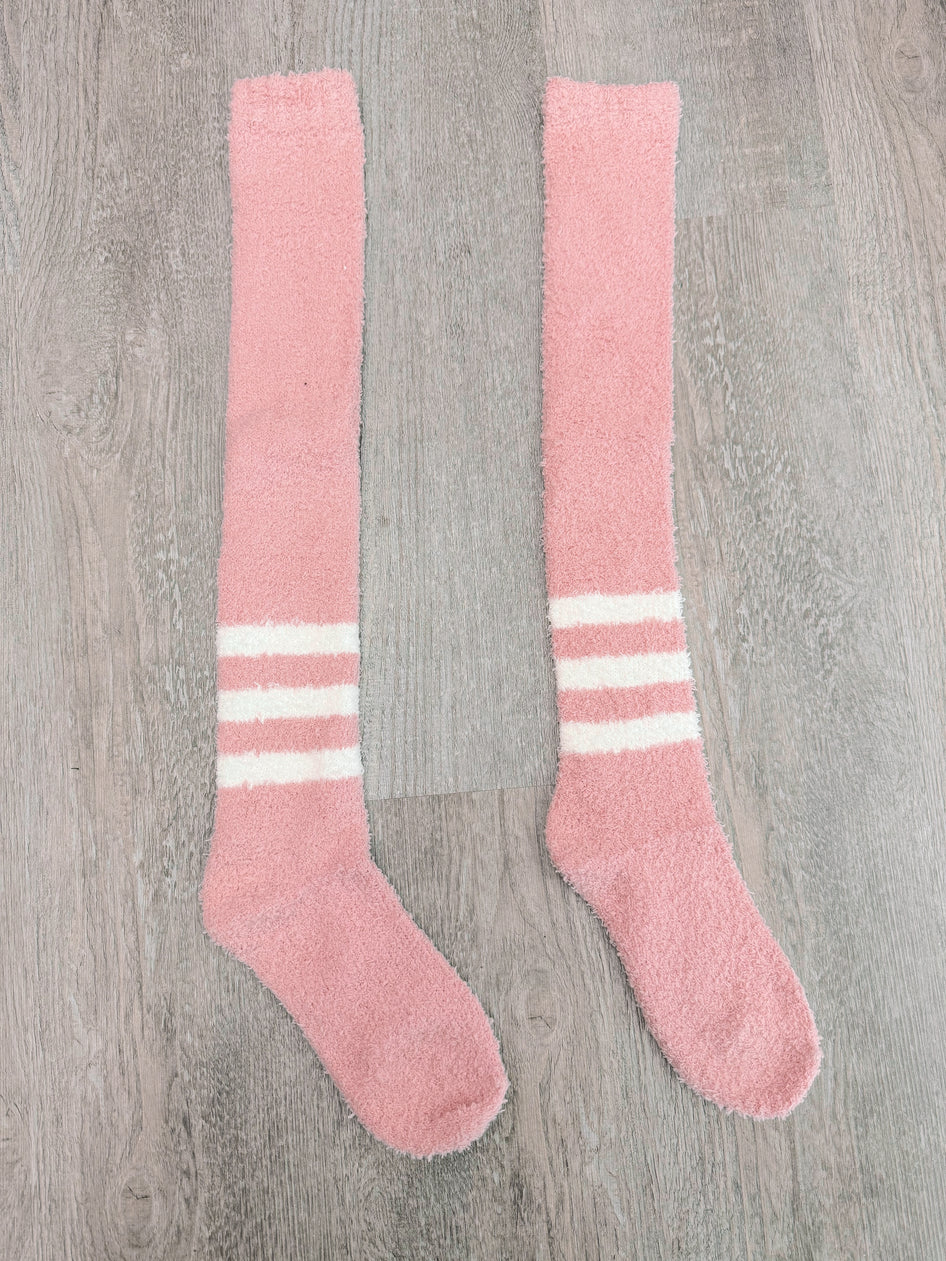 Pair of fluffy pink knee-high socks with white stripes, displayed on wood.